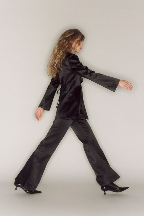 Person in a black satin suit (blazer and wide‑leg pants) walking forward against a neutral backdrop, showcasing motion and style.