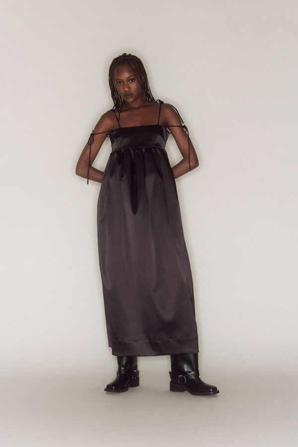 Model in a black satin maxi dress with thin straps, standing against a light background in black boots. Her arms are tied with ribbon-like accents for a chic look.