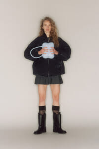 Young woman stands facing camera in a black oversized jacket, pleated skirt, and boots, holding a light-blue inflatable heart-shaped device with a tube.