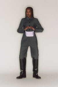 Full-body fashion shot of a model in a dark pinstripe suit with knee-high black boots, holding a small lavender handbag in front.