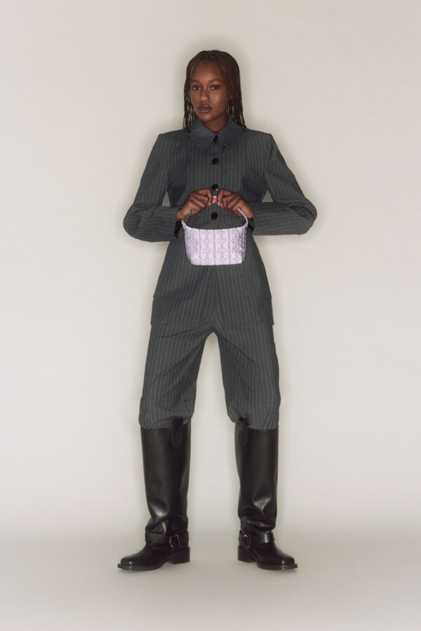 Full-body fashion shot of a model in a dark pinstripe suit with knee-high black boots, holding a small lavender handbag in front.