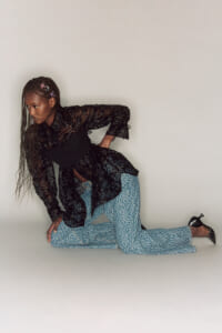 Woman with long braided hair wearing a black lace blouse and light blue patterned flare pants, posing in a studio on one knee with her hand resting on the ground.