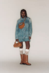 Person wearing an oversized denim jacket as a dress, tan crossbody bag, tan ankle boots, white socks, standing against a neutral backdrop.