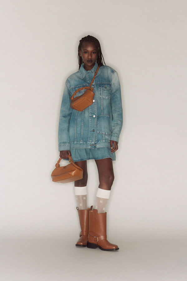 Person wearing an oversized denim jacket as a dress, tan crossbody bag, tan ankle boots, white socks, standing against a neutral backdrop.