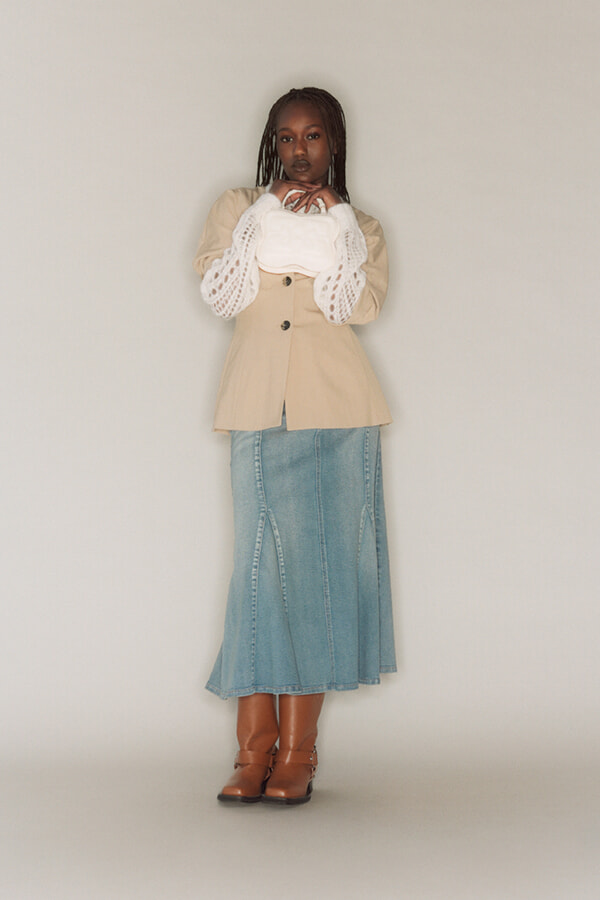 Full-length portrait of a woman wearing a beige blazer over a white lace-sleeve top, a blue denim midi skirt, and brown ankle boots, standing against a neutral background.
