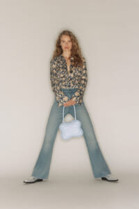 Woman standing in wide-legged jeans and a snake-print blouse, holding a light blue sculpted handbag against a plain backdrop.
