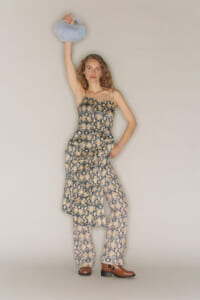Model wearing a snakeskin-print sleeveless dress with matching pants, standing with one arm raised holding a light blue plush bag above her head in a neutral studio setting.