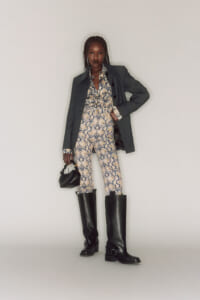 Full-length portrait of a woman wearing a snakeskin-patterned bodysuit with an oversized pinstripe blazer and black knee-high boots, holding a small black handbag.