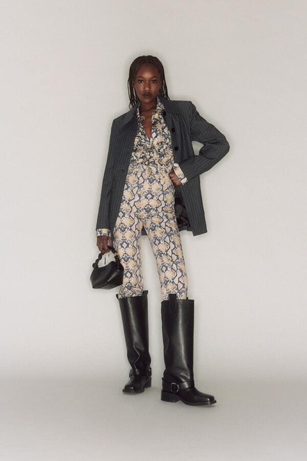 Full-length portrait of a woman wearing a snakeskin-patterned bodysuit with an oversized pinstripe blazer and black knee-high boots, holding a small black handbag.