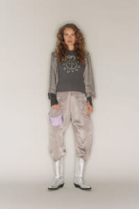 Young woman standing in a metallic satin outfit: oversized bomber jacket, matching pants, silver boots, and a small lavender handbag.