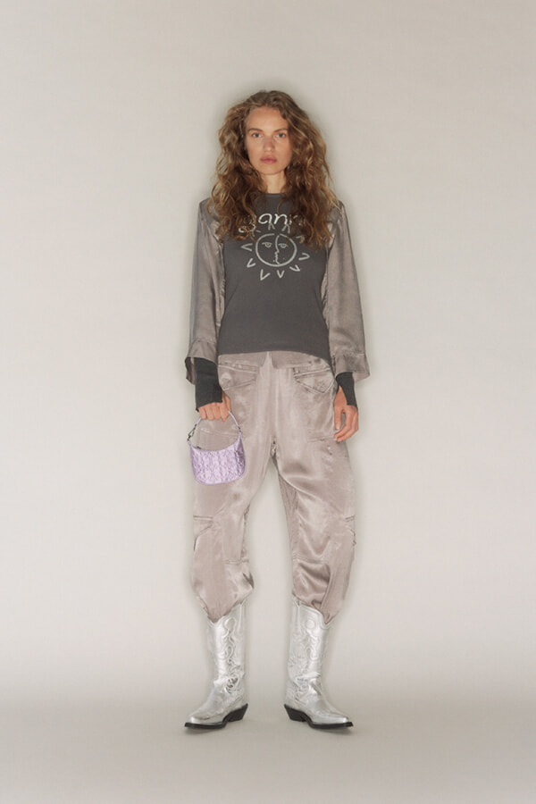 Young woman standing in a metallic satin outfit: oversized bomber jacket, matching pants, silver boots, and a small lavender handbag.