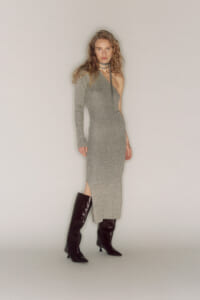 Model wearing a gray one-shoulder knit midi dress with a left sleeve and thigh-high slit, paired with black knee-high boots and a choker necklace.