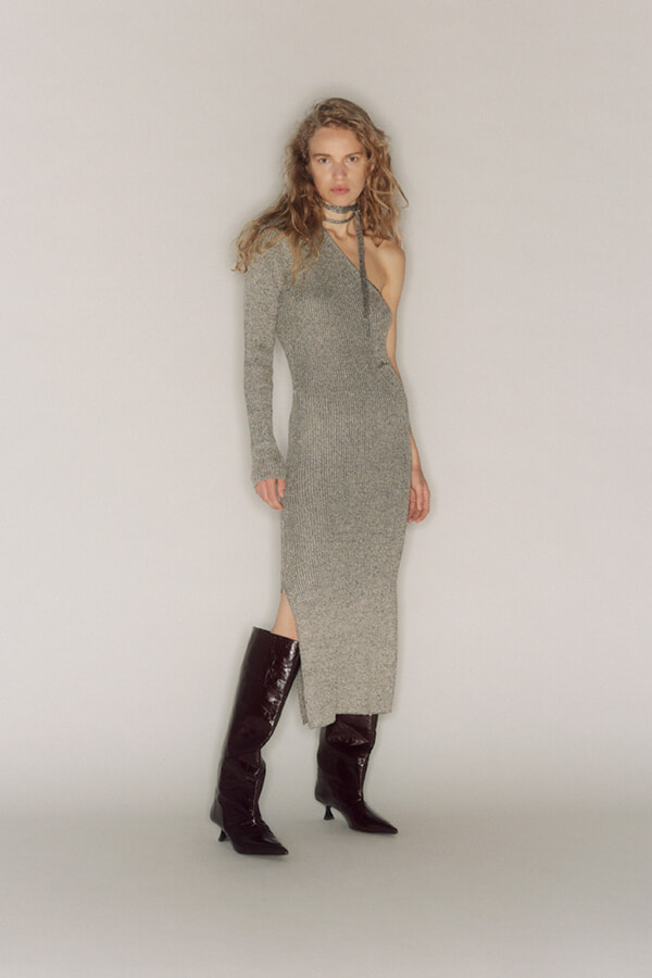 Model wearing a gray one-shoulder knit midi dress with a left sleeve and thigh-high slit, paired with black knee-high boots and a choker necklace.