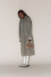 Woman in a long gray coat holding a small beige handbag, wearing metallic silver boots.