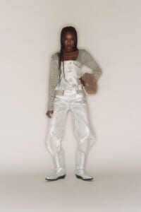 Model wearing a metallic white co-ord: cropped corset top with long-sleeve cardigan, high-waisted pants, white boots, and a beige fuzzy hat.