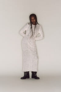 Woman wearing a long white sequin dress with black buckled boots, standing with hands on her hips against a neutral background.