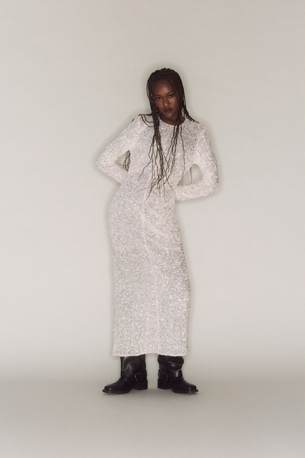 Woman wearing a long white sequin dress with black buckled boots, standing with hands on her hips against a neutral background.