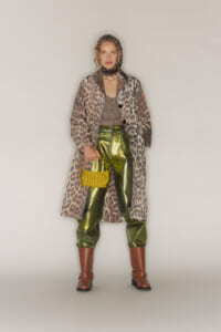 Model wearing a leopard-print long coat over a taupe top and shiny olive-green pants, with brown boots and a bright yellow knit handbag.