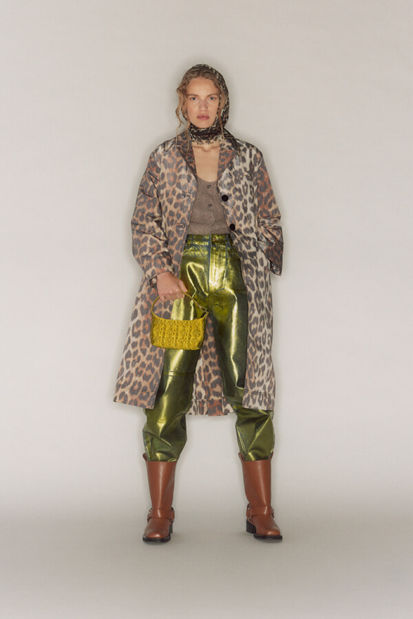 Model wearing a leopard-print long coat over a taupe top and shiny olive-green pants, with brown boots and a bright yellow knit handbag.
