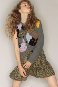 Woman with long wavy hair wearing a gray argyle cardigan over a patchwork sweater and a yellow plaid miniskirt, posed in a dynamic stance