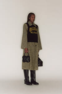 Woman standing in a studio wearing a checkered coat with a black knit vest that has a yellow GANNI logo, holding two black handbags, and wearing black knee-high boots.