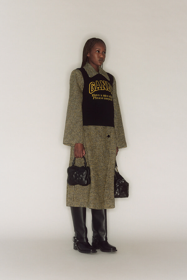 Woman standing in a studio wearing a checkered coat with a black knit vest that has a yellow GANNI logo, holding two black handbags, and wearing black knee-high boots.