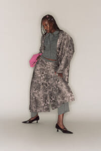 Person wearing a long gray floral coat over a teal outfit, holding a pink clutch and wearing black heels.