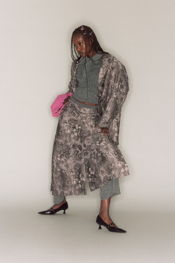 Person wearing a long gray floral coat over a teal outfit, holding a pink clutch and wearing black heels.