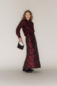Model wearing a burgundy lace sweater and matching floral lace maxi skirt, holding a small black handbag.