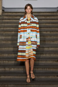 Model wearing a long, multicolored striped coat on stairs, paired with strappy sandals, facing forward.