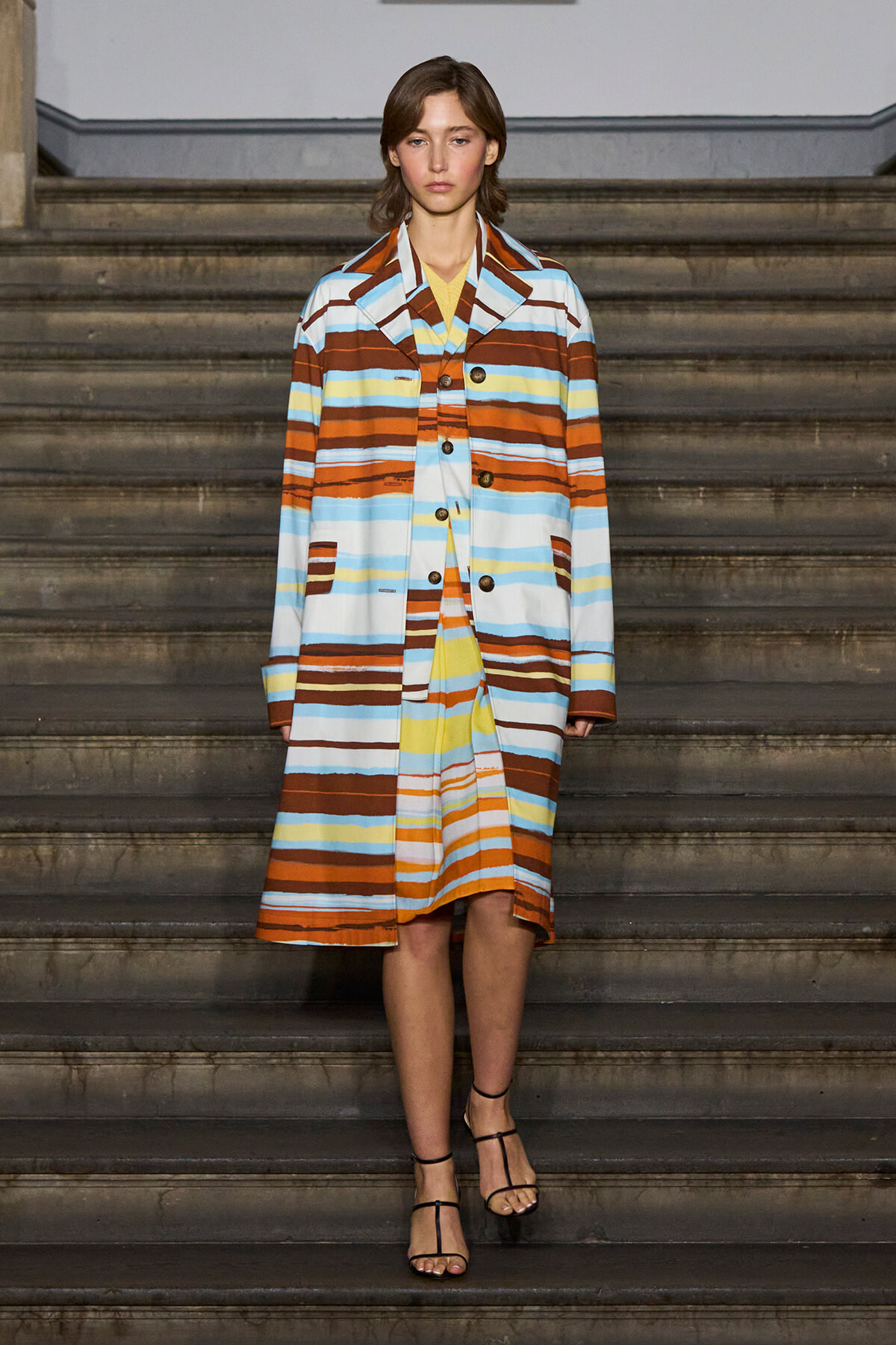 Model wearing a long, multicolored striped coat on stairs, paired with strappy sandals, facing forward.