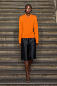 Model wearing a bright orange knit sweater and black wide knee-length shorts standing on stone steps.