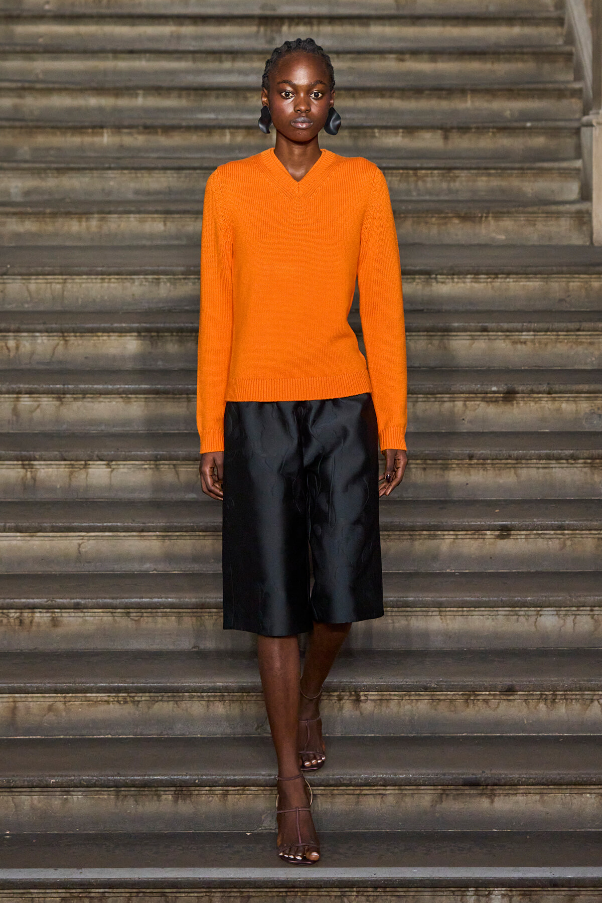 Model wearing a bright orange knit sweater and black wide knee-length shorts standing on stone steps.