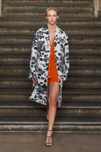 Model walking down a stone staircase in a white floral-print coat over an orange knit dress with black strappy heels, on a fashion runway backdrop.