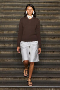 Model walking down stairs in a brown knit sweater, white knee-length shorts, black sandals, and large white statement jewelry in a fashion look.