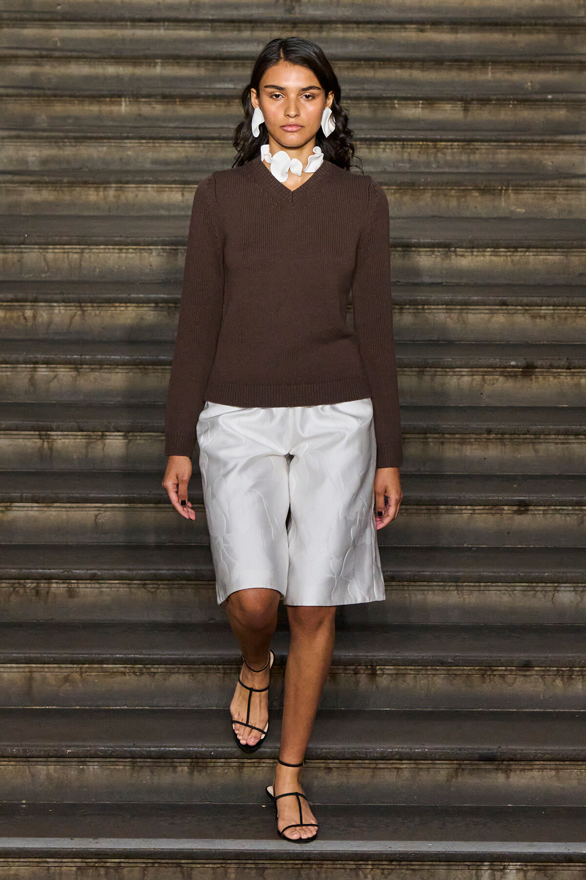 Model walking down stairs in a brown knit sweater, white knee-length shorts, black sandals, and large white statement jewelry in a fashion look.