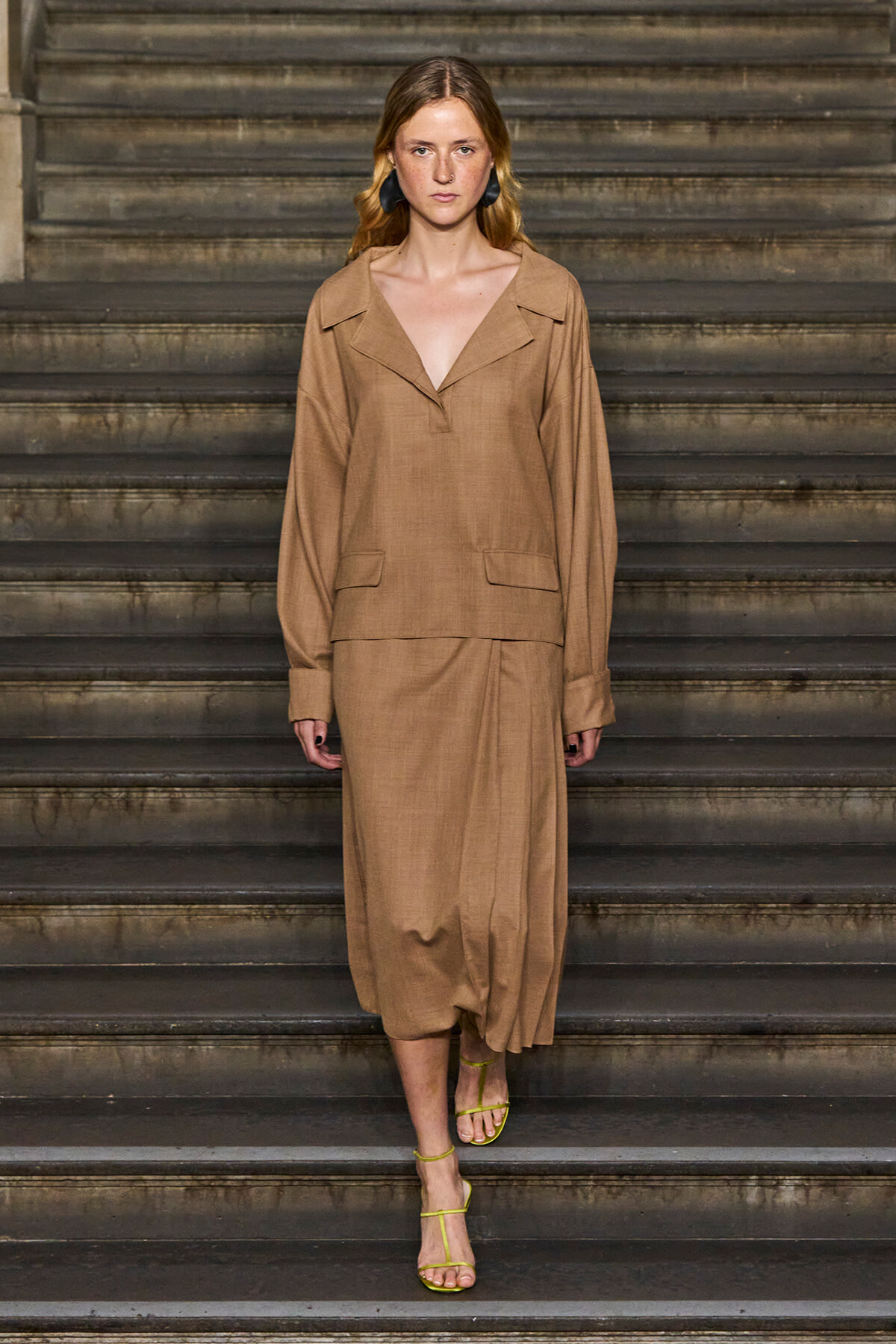 Full-body shot of a woman in a tan linen-style coordinated outfit (loose top with a wide collar and matching skirt) standing on a flight of dark stairs, wearing lime-green strappy sandals and black earrings.