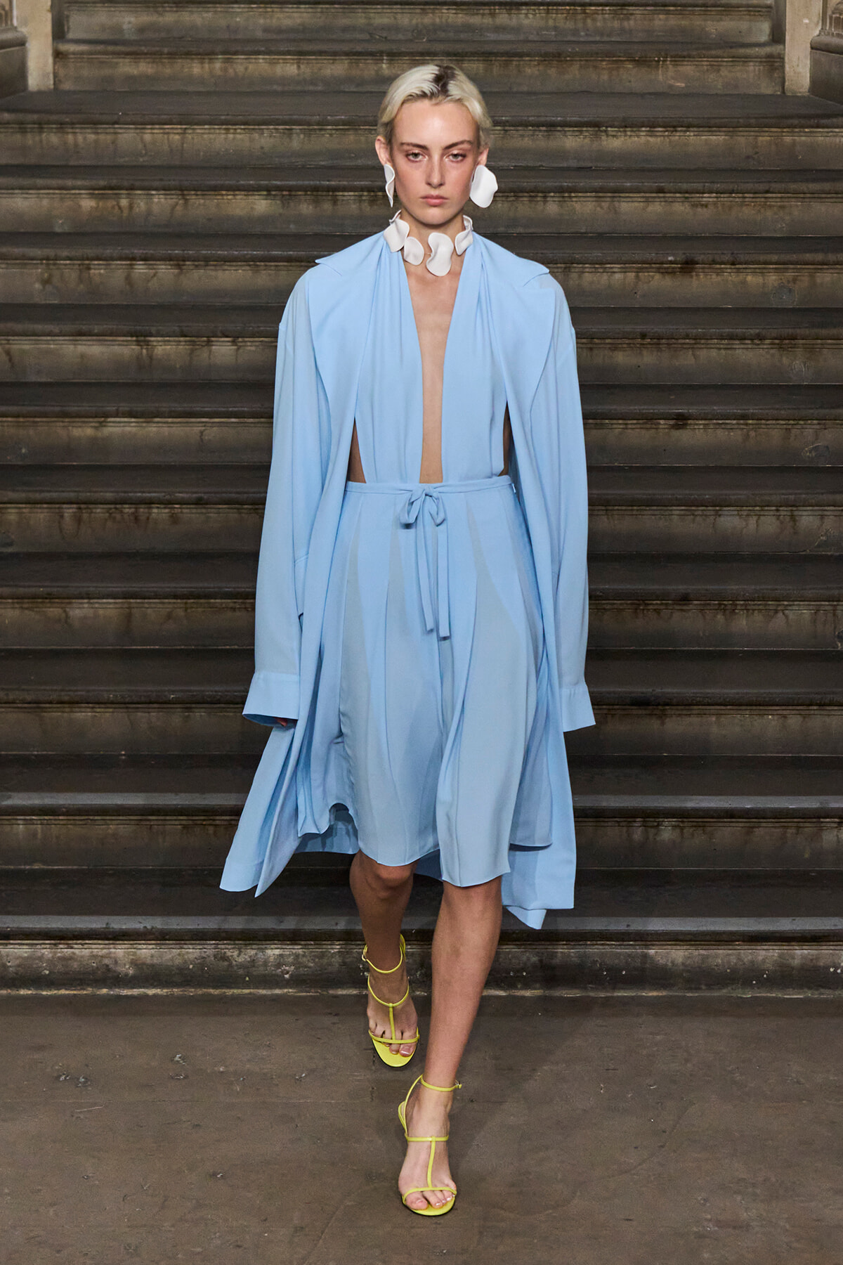 Model walking down stairs in a light blue, open-front dress with cape sleeves, paired with bright yellow strappy heels.