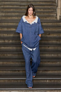 Model in a loose blue denim jumpsuit with a white collar walks down a flight of stairs, hands in pockets.