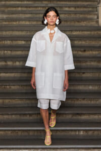 Female model on a runway wearing a sheer white oversized shirt with large front pockets, white shorts, and bright yellow strappy sandals.