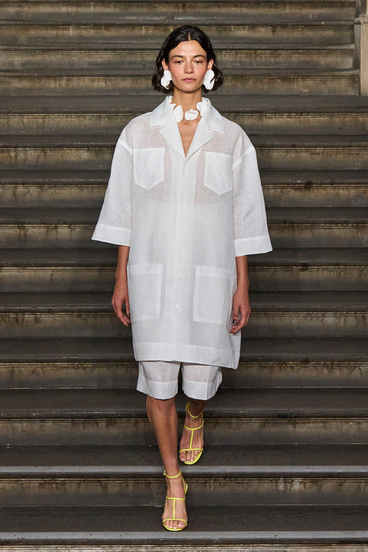 Female model on a runway wearing a sheer white oversized shirt with large front pockets, white shorts, and bright yellow strappy sandals.