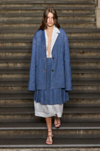 Model wearing an oversized blue denim blazer and matching pleated skirt over a white dress, standing on stone stair steps.