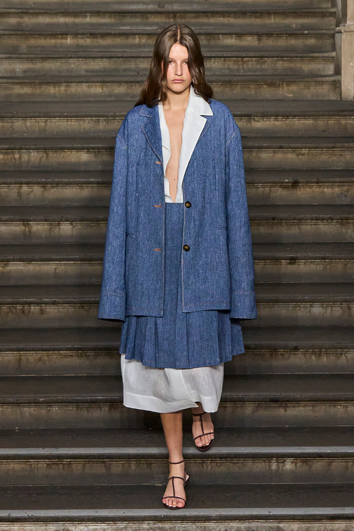 Model wearing an oversized blue denim blazer and matching pleated skirt over a white dress, standing on stone stair steps.