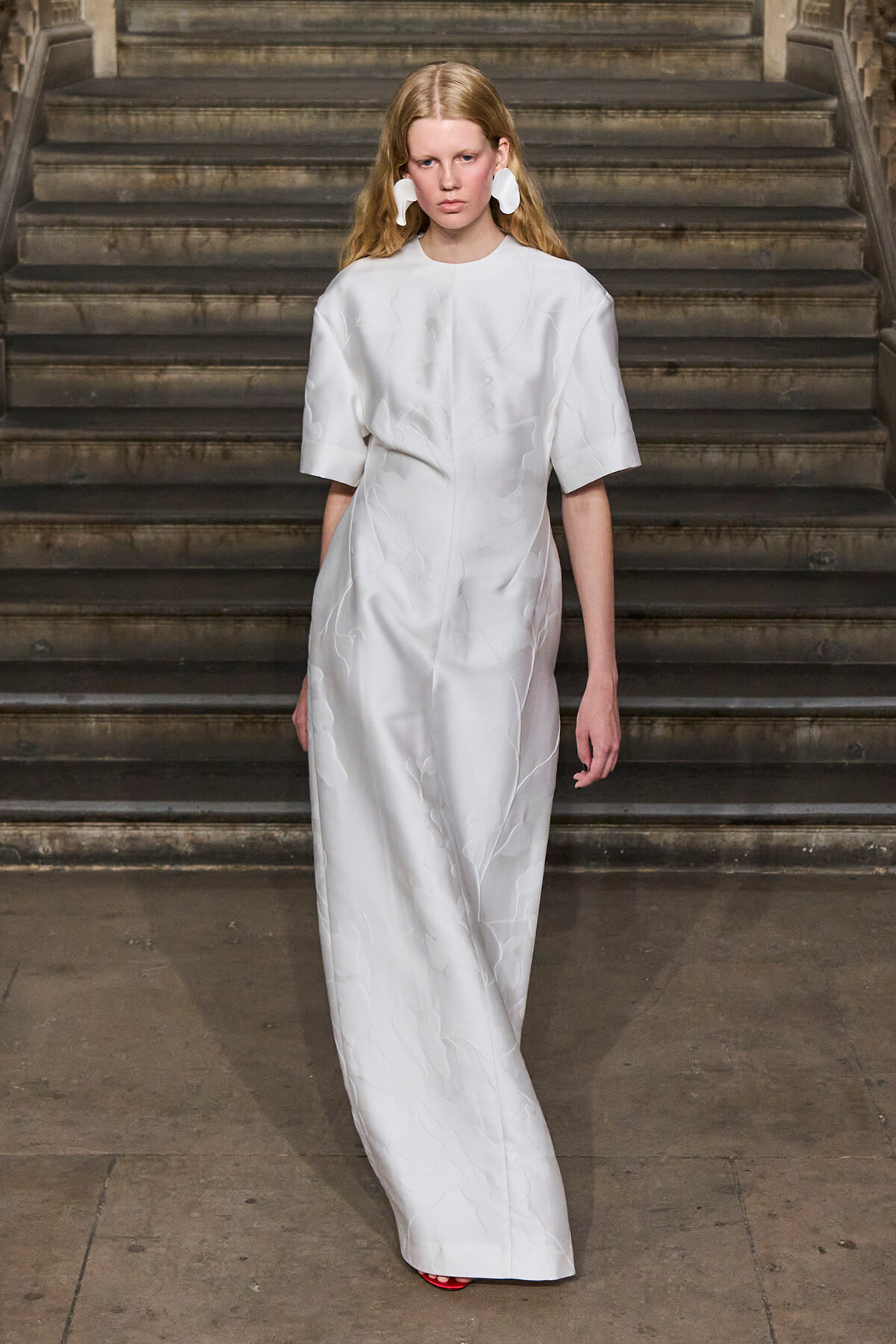 Female model wearing a white textured, midi-length dress with wide sleeves on a staircase runway.