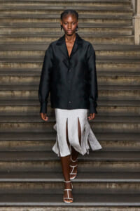 Model walking down a stone staircase in a black satin oversized blazer and a white split-front skirt with flowing panels, wearing white strappy heels.