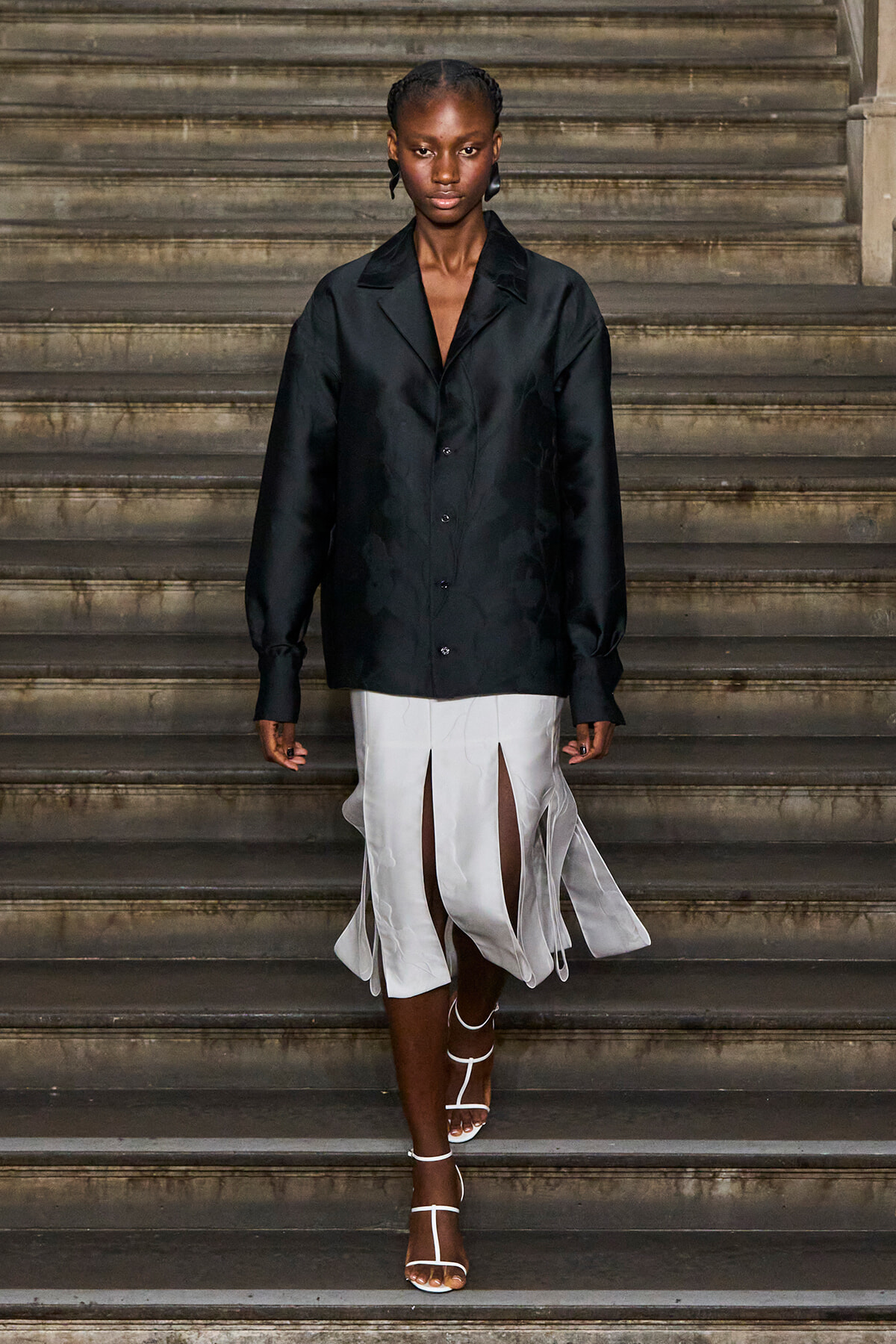 Model walking down a stone staircase in a black satin oversized blazer and a white split-front skirt with flowing panels, wearing white strappy heels.