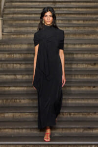 Woman in a long black dress with a twisted knot at the chest, walking down stone steps in red strappy sandals.