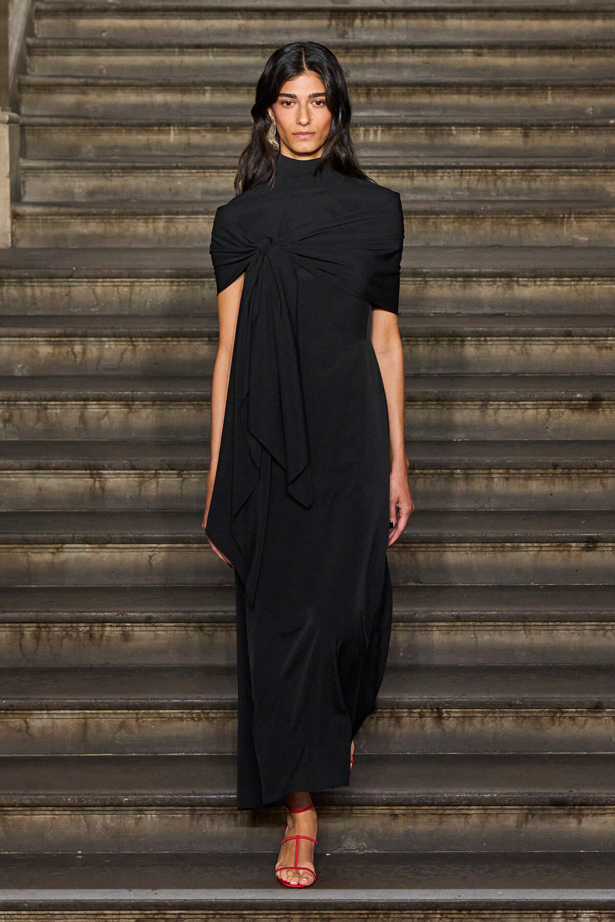 Woman in a long black dress with a twisted knot at the chest, walking down stone steps in red strappy sandals.