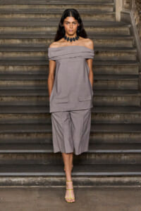 Woman wearing an off-the-shoulder gray pantsuit stands on stone stairs, accessorized with a bold black necklace and lime-green sandals.