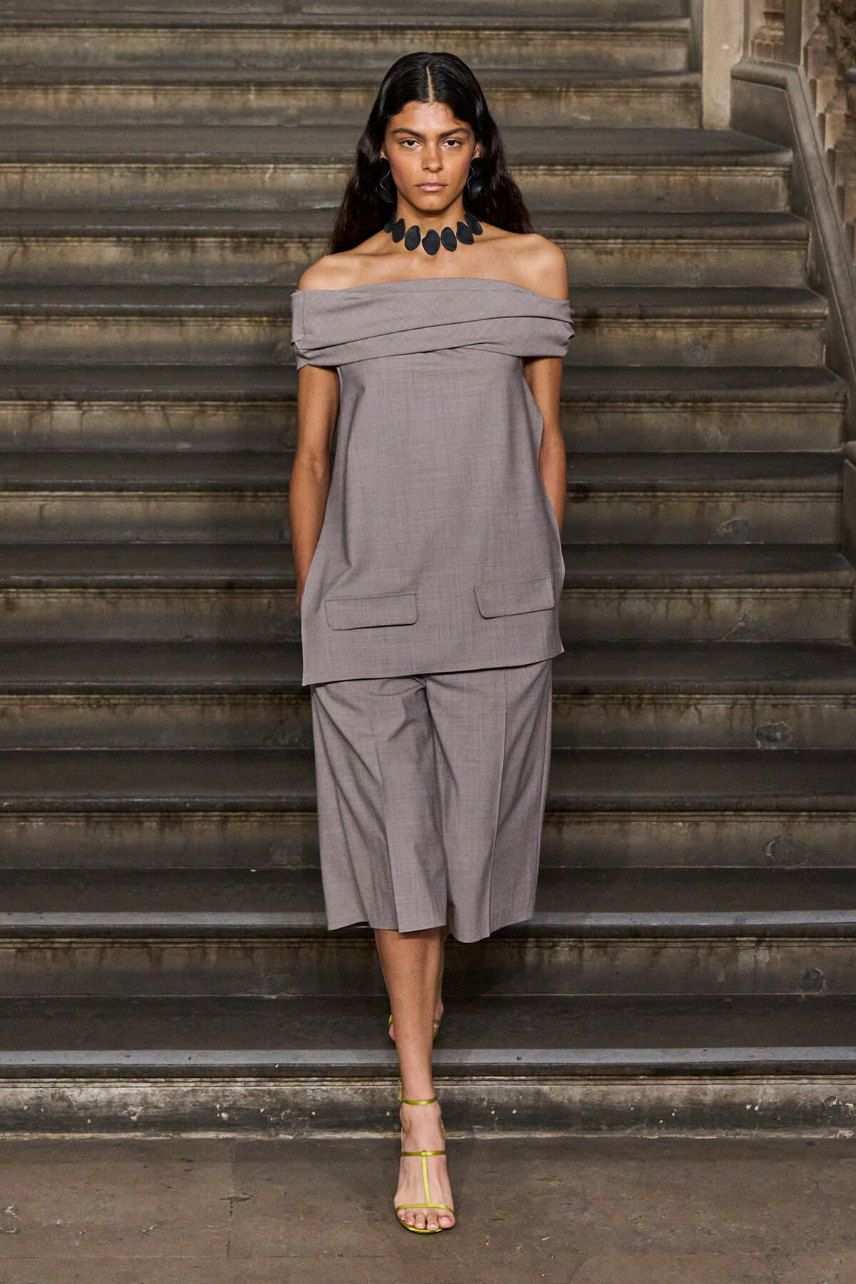 Woman wearing an off-the-shoulder gray pantsuit stands on stone stairs, accessorized with a bold black necklace and lime-green sandals.